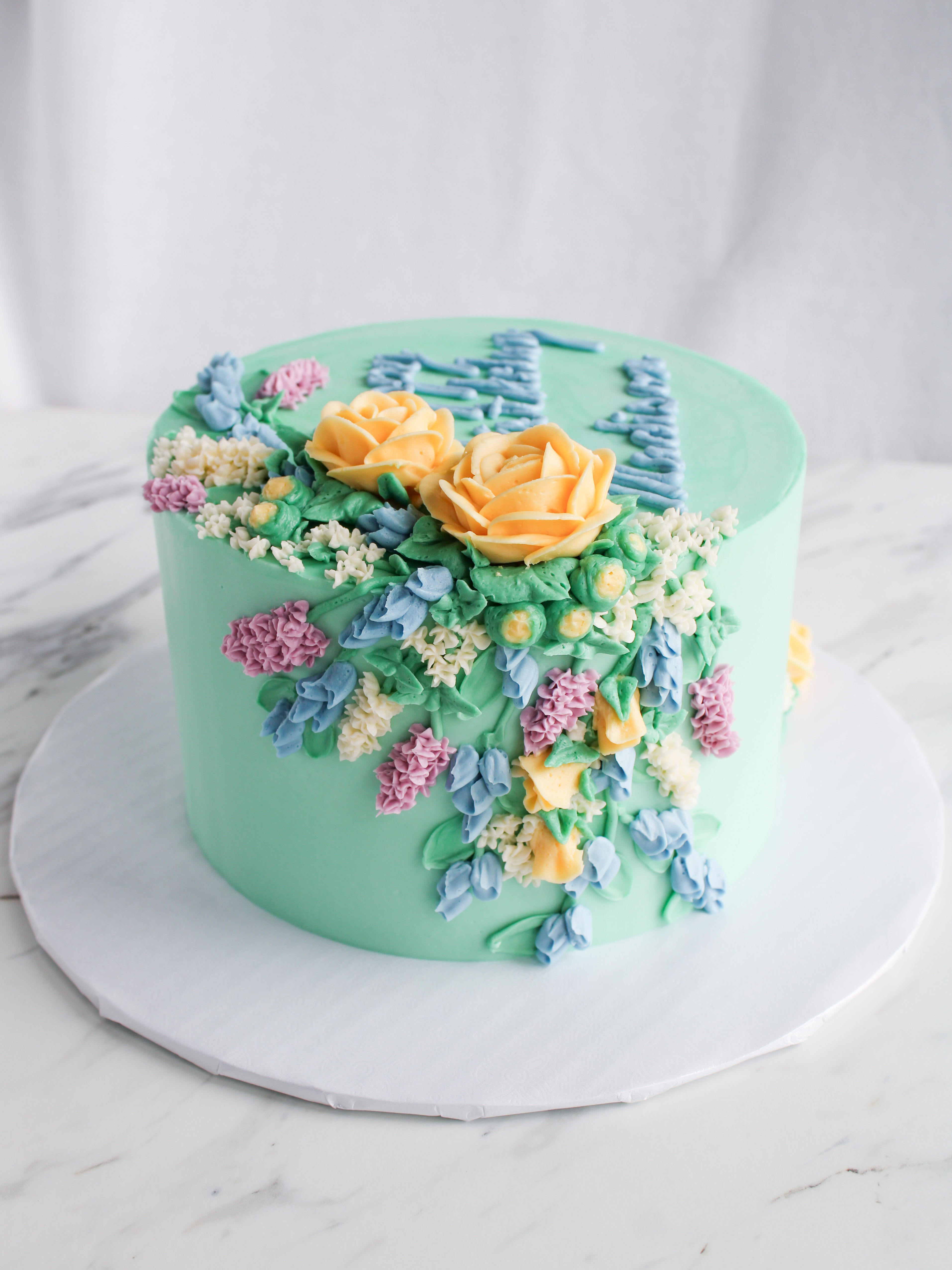 buttercream floral cake with yellow roses purple lilacs and blue delphinium